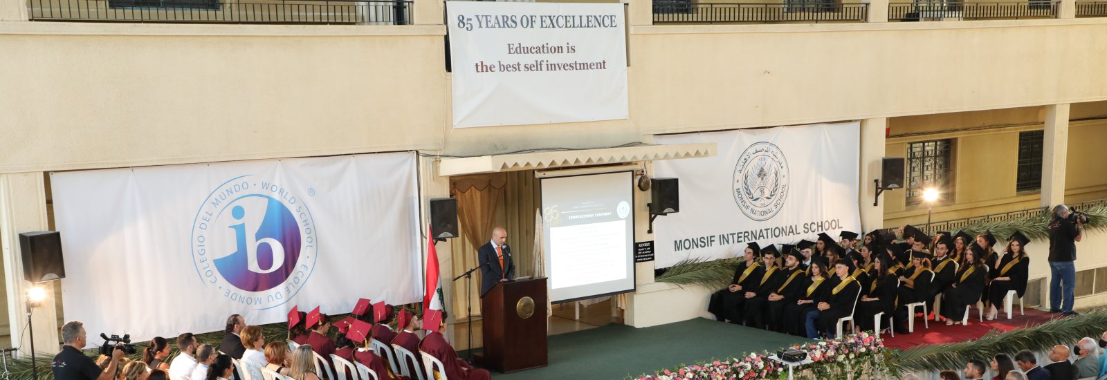 Commencement ceremony in the school courtyard: graduates in black and maroon gowns seated in front of banners reading 85 Years of Excellence, the IB World School seal, and Monsif International School.